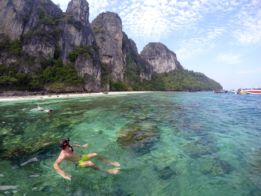 A man snorkeling in clear seawater with towering limestone cliffs along the shoreline of Mosquito Island in Koh Phi Phi