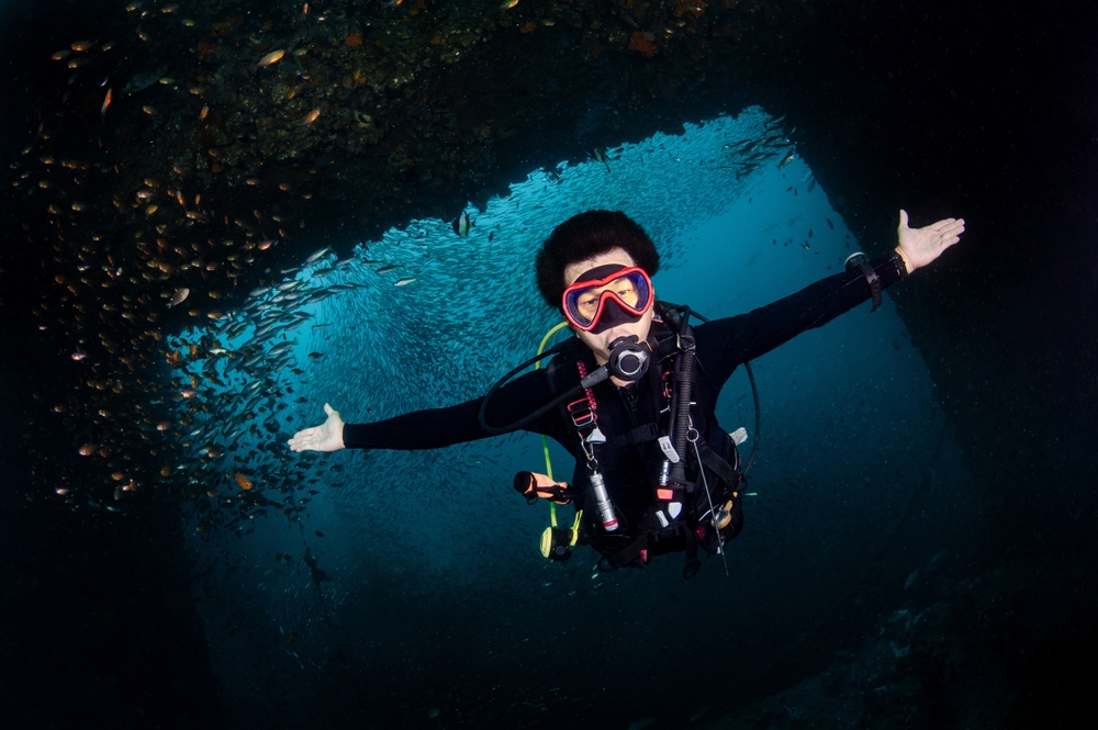 A diver underwater with a huge school of fish swimming in the background at King Cruiser Wreck in Koh Phi Phi