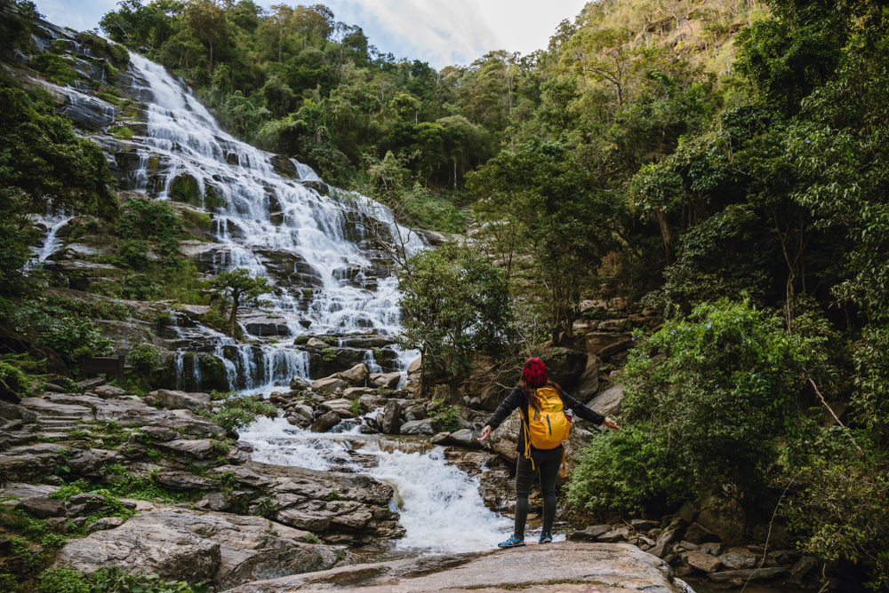 A tourist with a yellow backpack admiring Mae Ya Waterfall, a spectacular multi-tiered waterfall in Thailand surrounded by lush green forest.