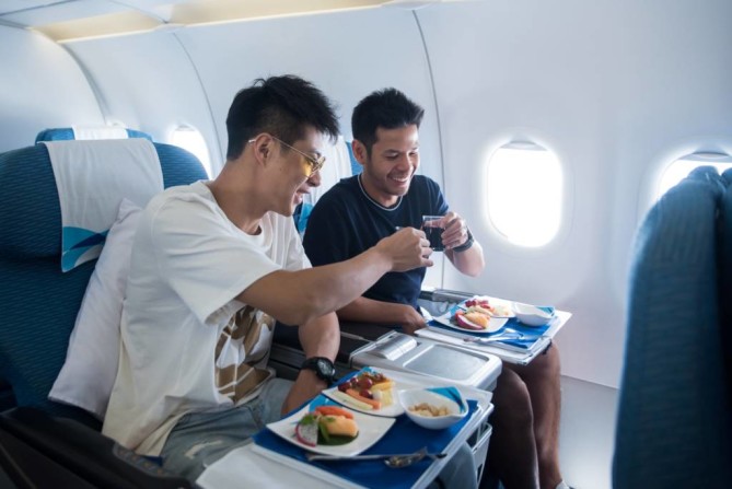 Two male passengers smiling and toasting drinks while enjoying an in-flight meal.
