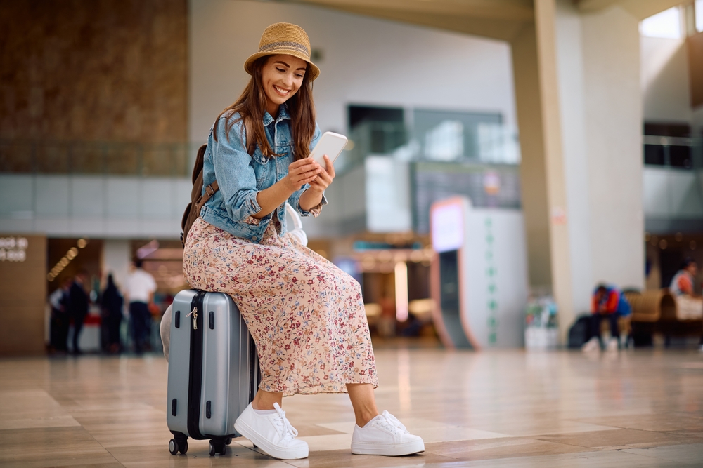 Female tourist sitting on a suitcase checking her TDAC confirmation email on a smartphone.