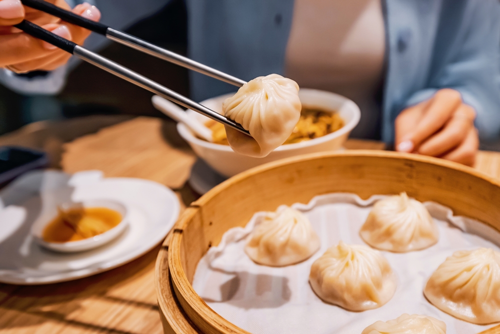  Chopsticks picking up a fresh Xiaolongbao steamed soup dumpling from a bamboo basket, a famous food in Shanghai.