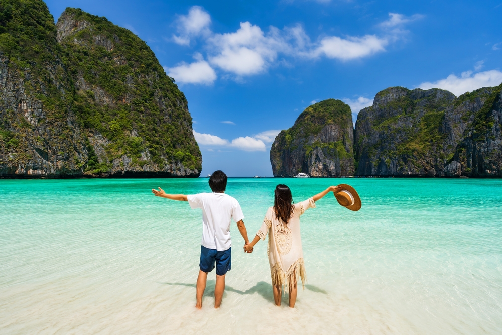 คู่รักยืนจับมือริมหาดทรายขาวหันหน้ามองวิวรอบอ่าวมาหยา กระบี่ อย่างมีความสุข