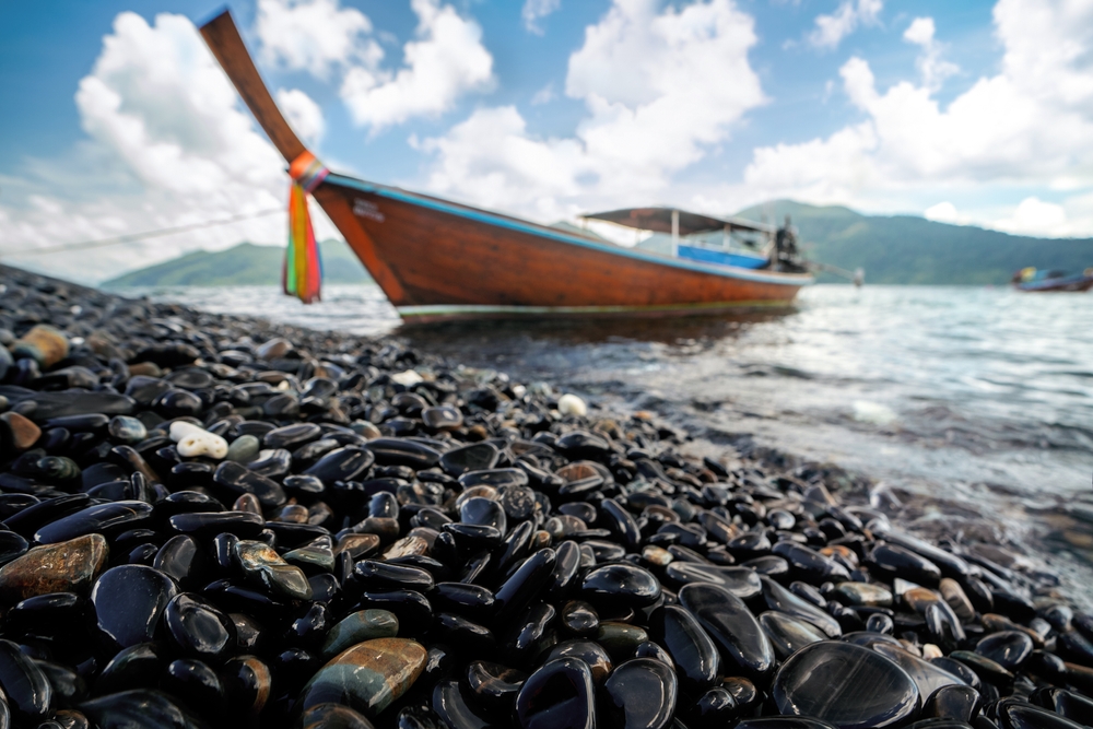 หินสีดำมันวาวเรียงรายริมชายหาดตัดกับวิวเรือหางยาวที่เกาะหินงาม สตูล ความงามธรรมชาติที่สายเที่ยวคนเดียวห้ามพลาด